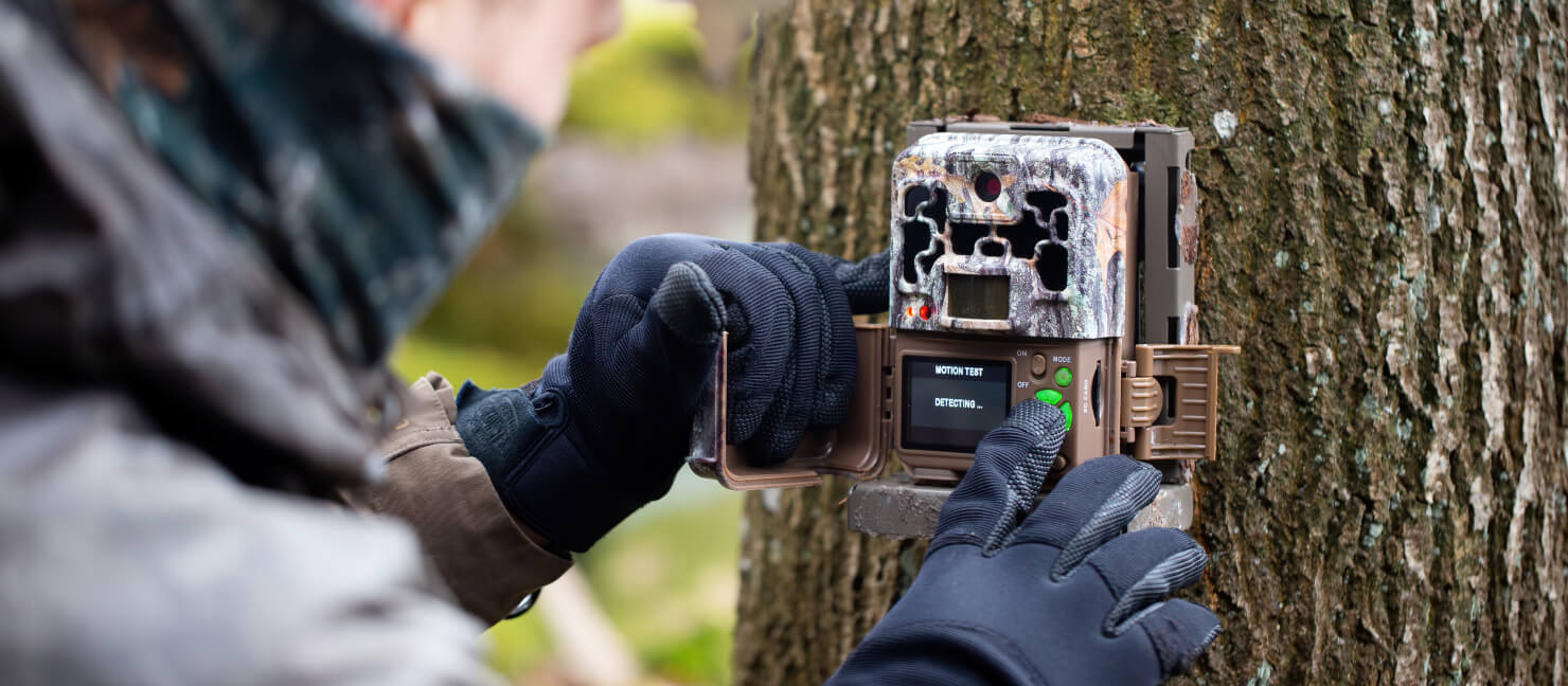 A hunter wears black gloves and sets up his game camera, which is secured to a tree.