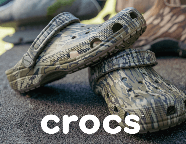 Pair of camouflage-pattern Crocs clogs displayed outdoors on a weathered wooden surface.