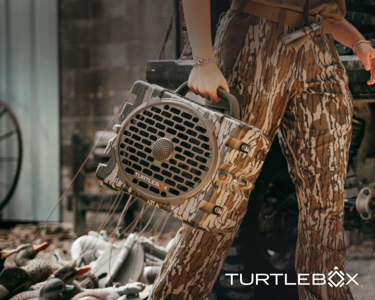 Camouflage Turtlebox portable speaker carried outdoors, with TURTLEBOX logo.