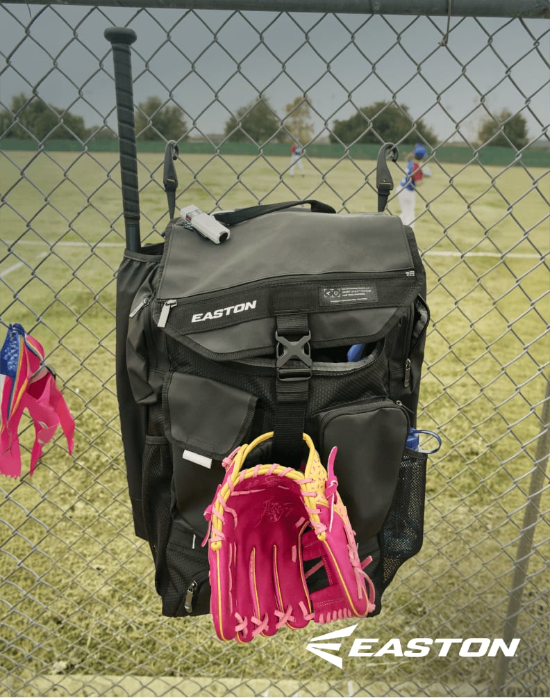 Easton baseball backpack hanging on a chain link fence with pink and yellow batting gloves tucked into exterior pocket.