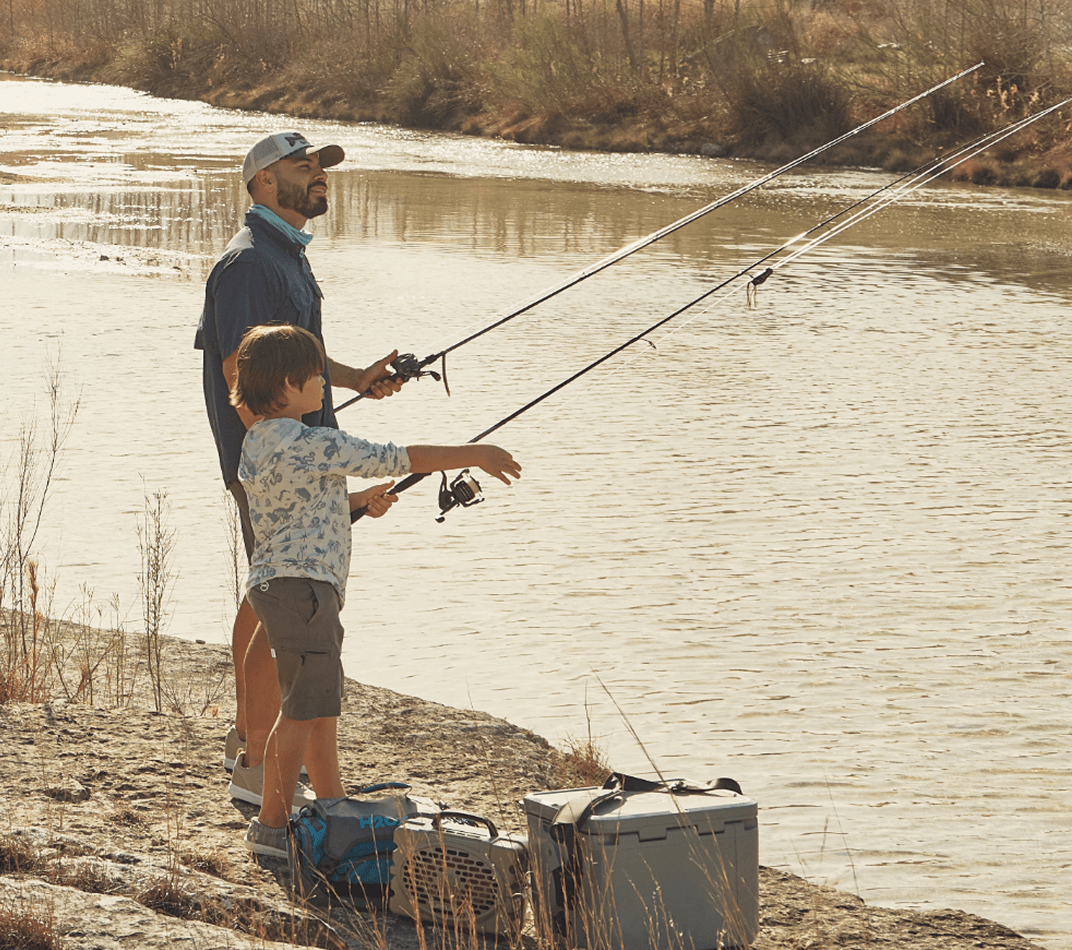 Two people fishing from the edge of a lake, standing beside a tackle box and cooler.