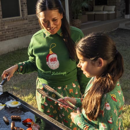 Adult and child wearing matching green Magellan Outdoors holiday apparel cook on an outdoor griddle.