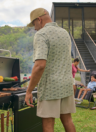 Outdoor lifestyle scene showing a person grilling by a lake, wearing a BURLEBO short-sleeve patterned shirt and shorts.