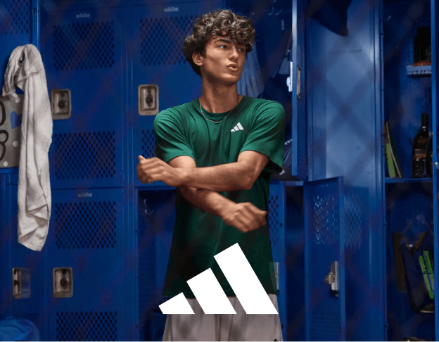 Person in a green adidas shirt standing in front of blue lockers with sports apparel hanging nearby.