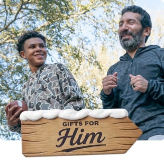 Person holding a football walks outdoors beside another person in athletic apparel. A wooden holiday sign with snow reads “Gifts for Him.”