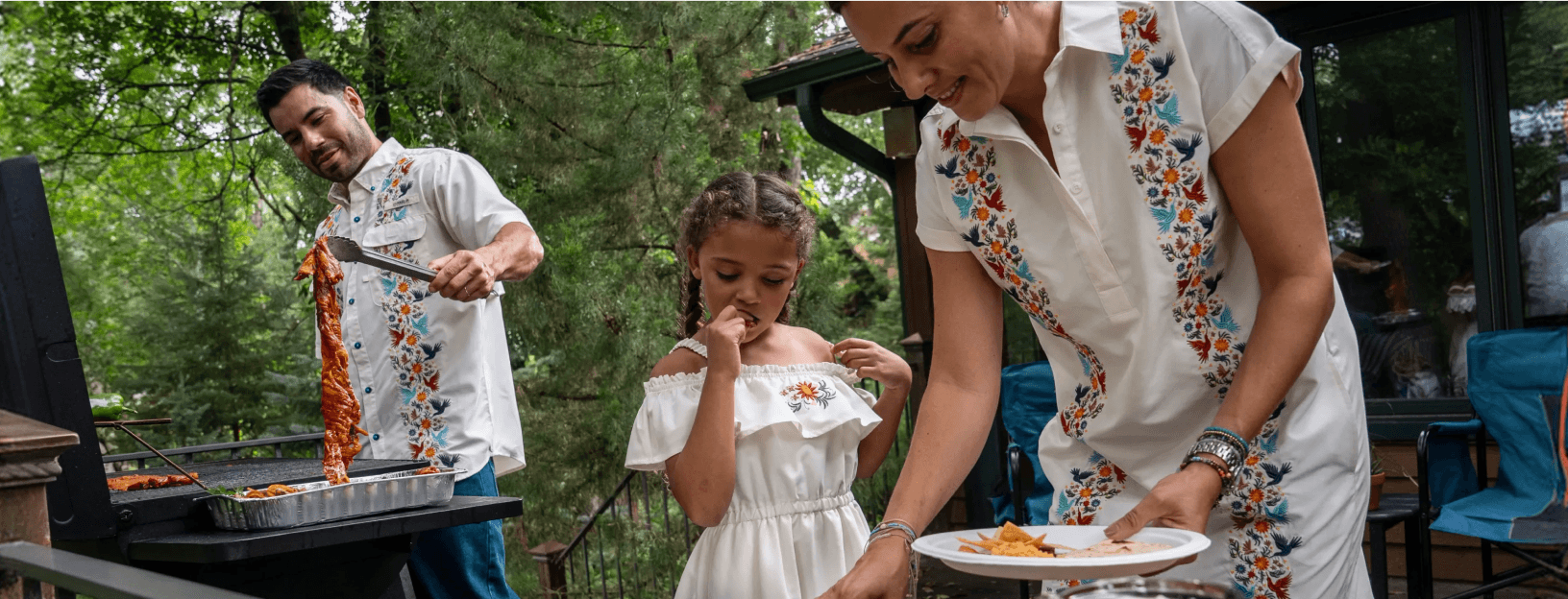 Person grilling ribs on an outdoor smoker while another person serves food beside a child standing on a backyard patio surrounded by trees. All three wear matching Magellan Outdoors white Hispanic Heritage shirts or dresses with floral embroidery.