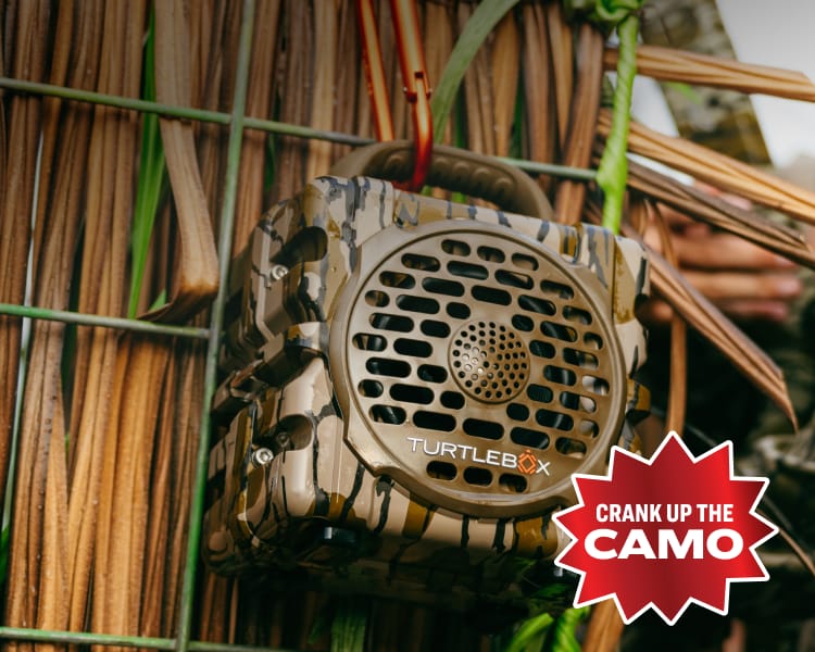Close-up of a Turtlebox Original Gen 3 Mossy Oak Bottomland speaker mounted outdoors among camouflage-patterned gear. Overlay red badge reading “Crank Up the Camo.”