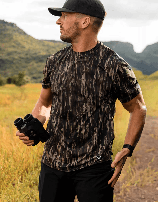 Person wearing a camouflage go-to t-shirt outdoors in rugged landscape holding gear.