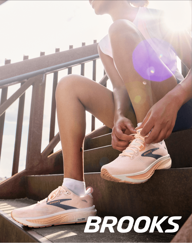 Runner seated on outdoor steps tying Brooks running shoes in bright sunlight.