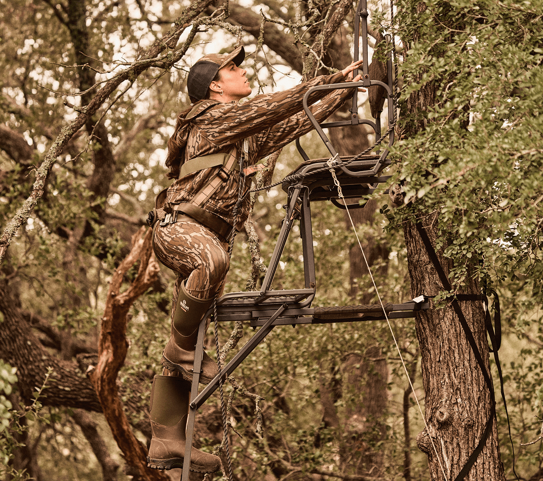 Macy Watkins Johnston wearing Magellan Outdoors Fayette Collection camouflage hunting apparel, a safety harness, and rubber boots climbing into a tree stand attached to a tree in a wooded area.