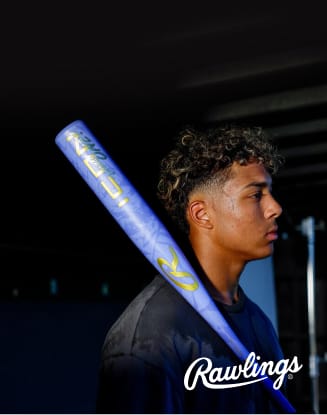 Person holding a blue Rawlings baseball bat over one shoulder against a dark indoor sports setting.