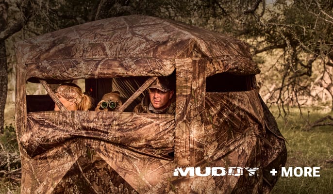 Hunters inside a camouflage hunting blind in a wooded setting.Muddy logo and “+ More” appear at the bottom of the image.