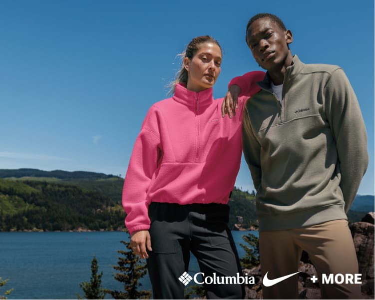 Two models wearing Columbia outdoor apparel, including a bright pink pullover and dark green jacket, standing outdoors against a blue sky.