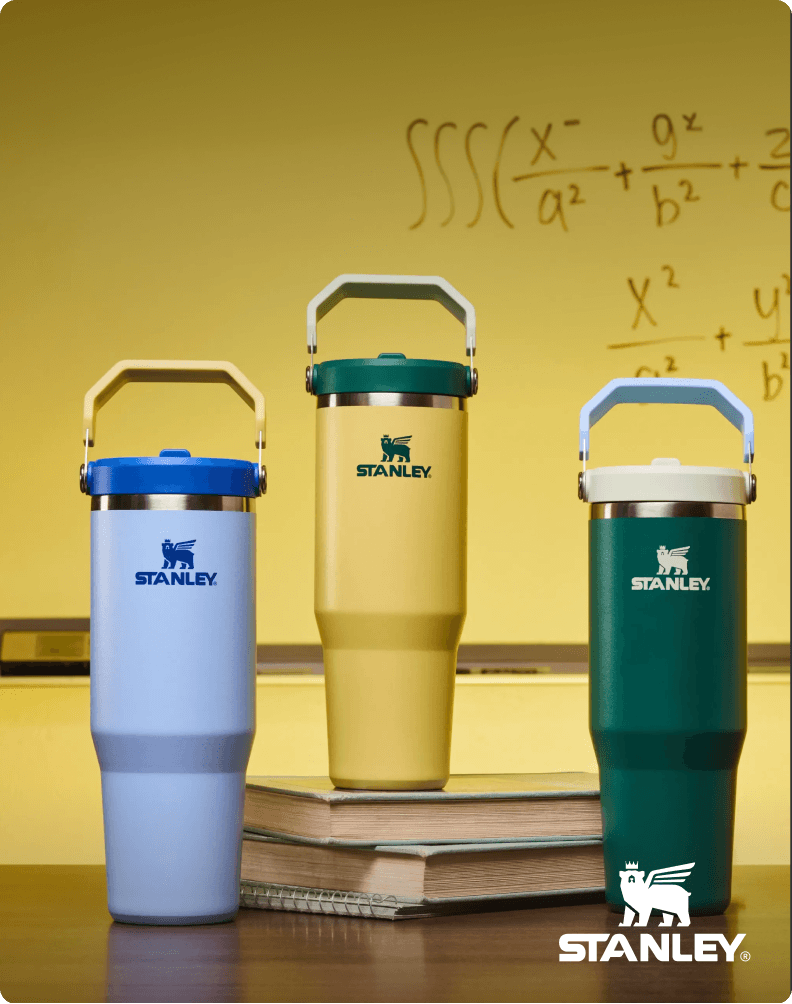 Three Stanley drink tumblers in blue, yellow, and green with handles are displayed in front of a classroom chalkboard with math equations, alongside Stanley branding.