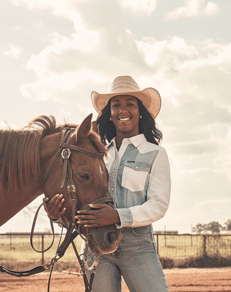 Person wearing a cowboy hat and denim outfit standing beside a saddled horse in an outdoor ranch setting.