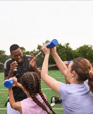 Young soccer athletes on a green soccer field spraying water from bottles onto a person in black athletic gear, with trees and soccer equipment in the background.