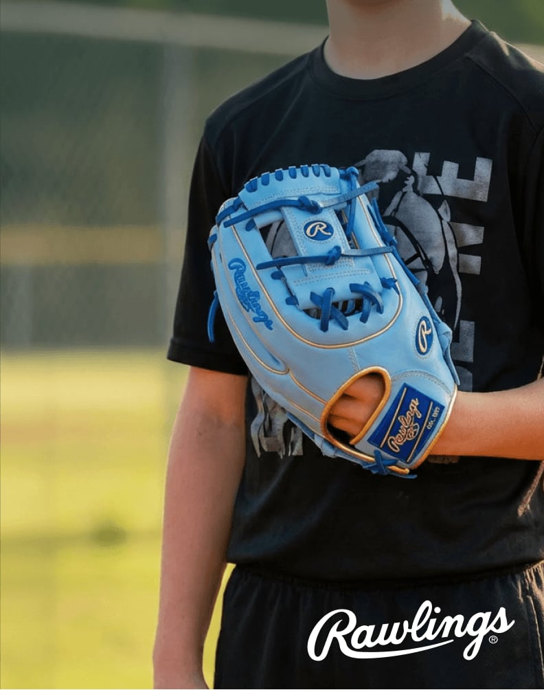 A person wearing a light blue Rawlings heart of the hide baseball glove while standing on a baseball field, shown from the shoulders down.
