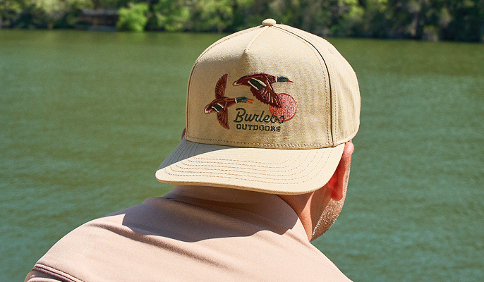 Close-up of a tan BURLEBO baseball cap with an embroidered outdoor logo, worn by a person facing a lake.