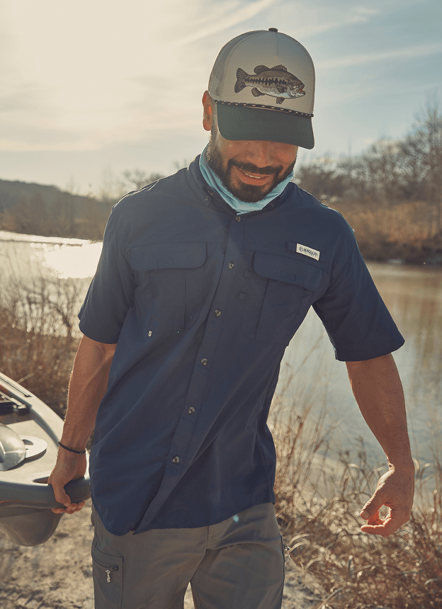 Person wearing a Magellan Outdoors dark short-sleeve fishing shirt and cap standing outdoors near tall grass.