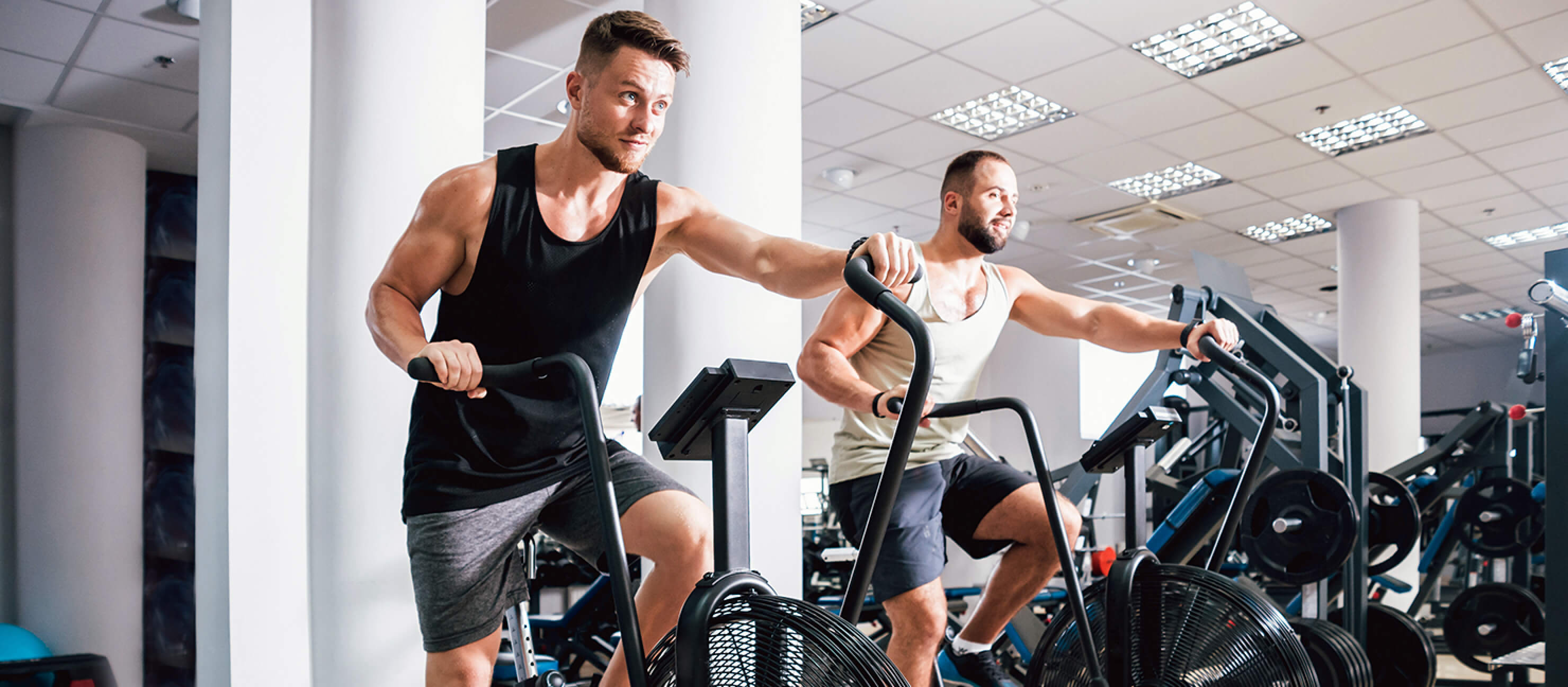 Two men at the gym pedal on dual action fan exercise bikes