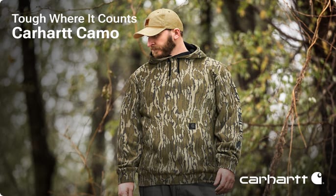 Person wearing a camouflage hooded jacket and camouflage cap standing outdoors among trees.