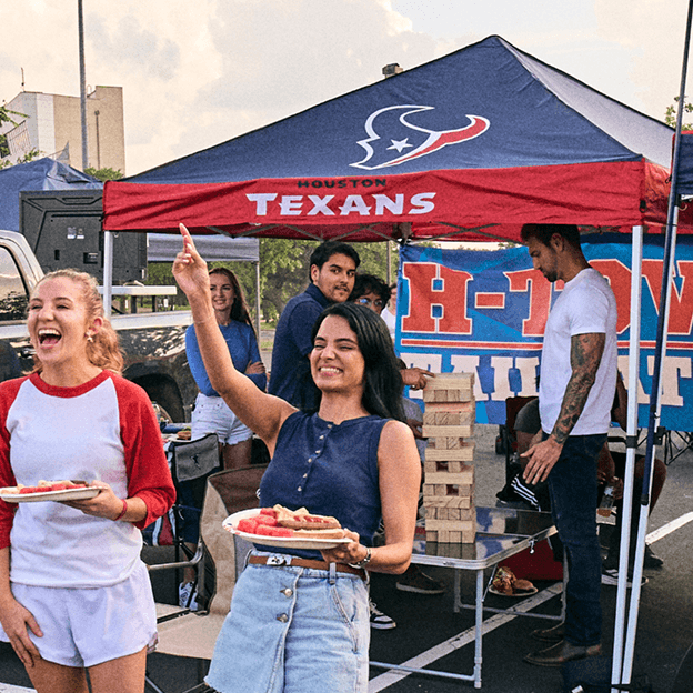 Fans gathered near a Houston Texans tailgating tent with team branding, food, and outdoor seating.