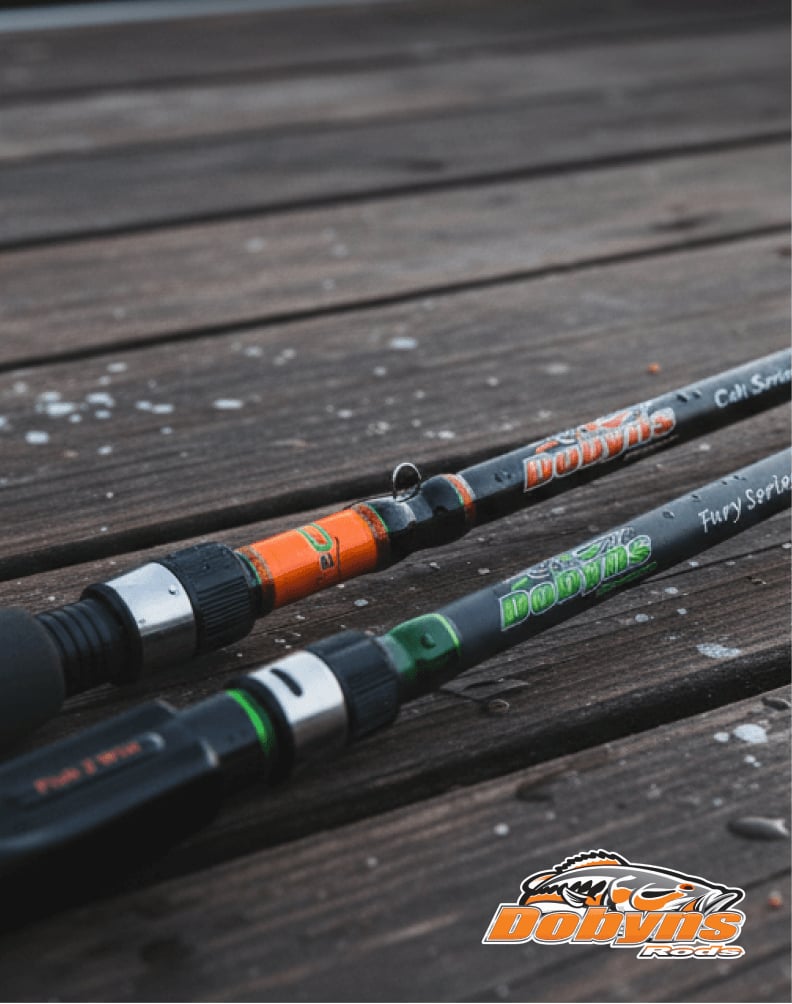 Two Dobyns fishing rods resting on a weathered wooden dock, one with orange accents and one with green accents, shown close‑up with visible branding.