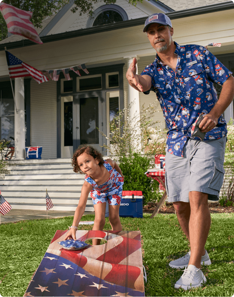 Adult playing cornhole with child in front yard decorated with American flags promoting patriotic summer clothing styles.