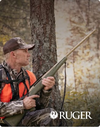Hunter in camouflage clothing holding a bolt-action rifle while sitting beside a tree in a wooded area.
