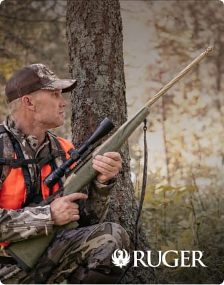 Hunter in camouflage clothing holding a bolt-action rifle while sitting beside a tree in a wooded area.