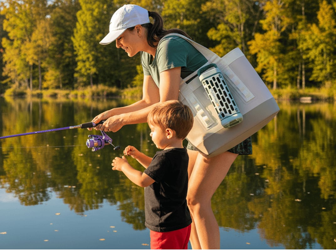 Adult and child fishing together at a calm lake surrounded by trees on a sunny day with a YETI tote and Turtlebox speaker.