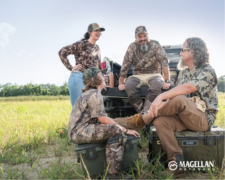 Four people wearing camouflage apparel gathered around the back of a vehicle in a grassy field.