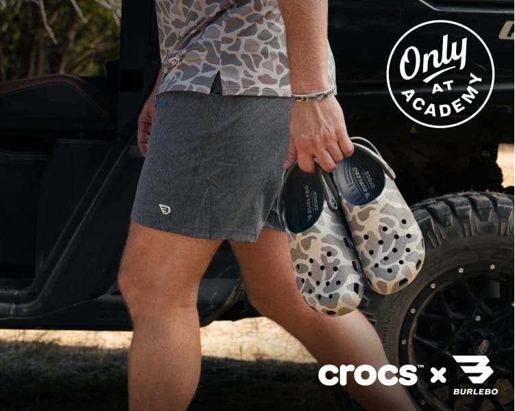 Person carrying camouflage-pattern Crocs footwear while walking beside an off-road vehicle.