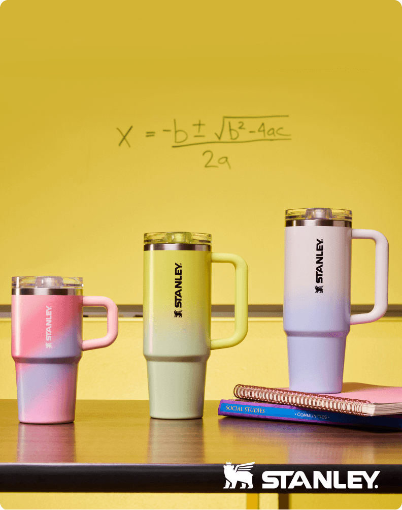 Three Stanley drink tumblers in blue, yellow, and green with handles are displayed in front of a classroom chalkboard with math equations, alongside Stanley branding.