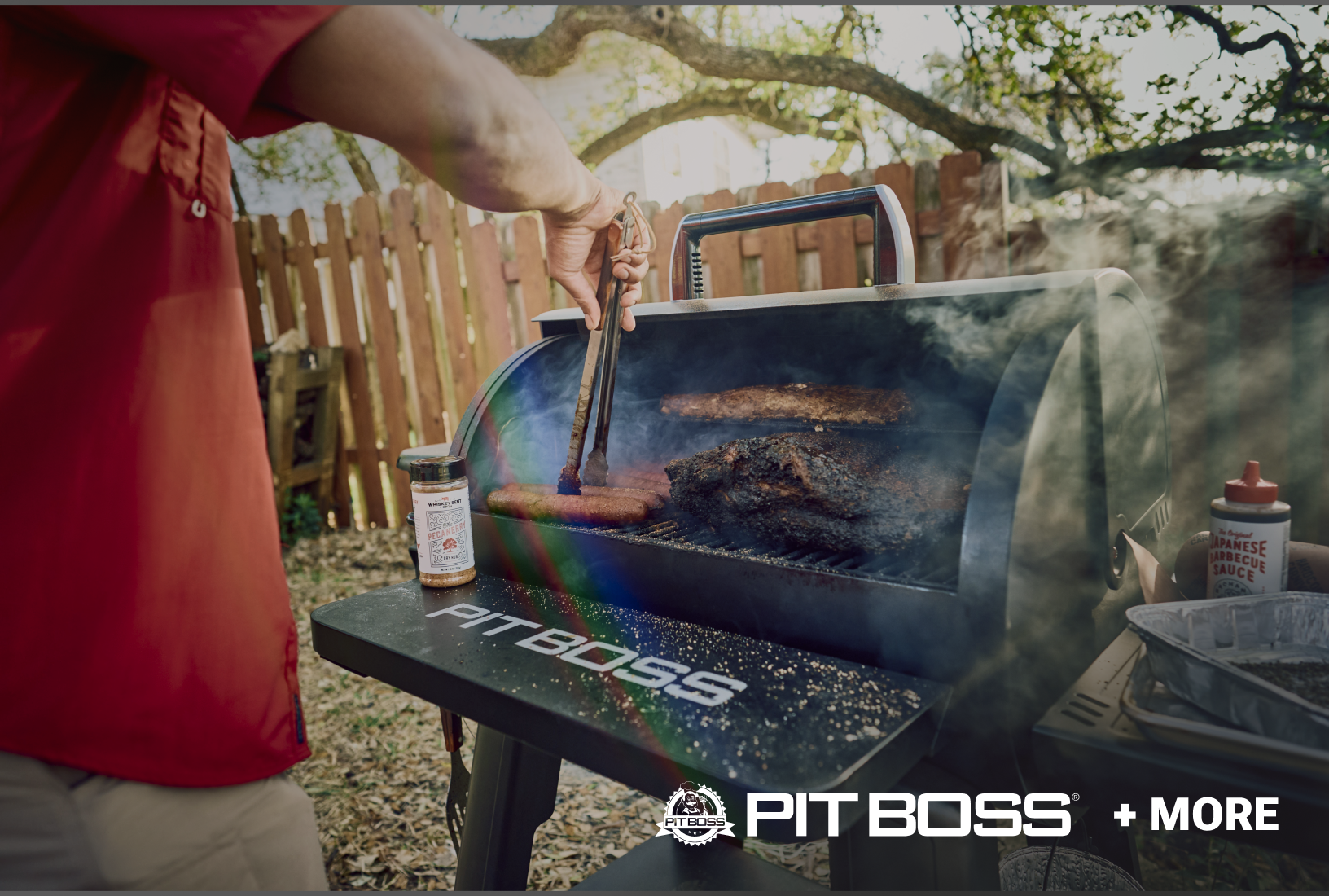 Close-up of a smoker grill cooking a large piece of meat and sausages with smoke rising, a hand using tongs to turn food, seasoning on the side shelf labeled “PIT BOSS,” and text “PIT BOSS + MORE.”