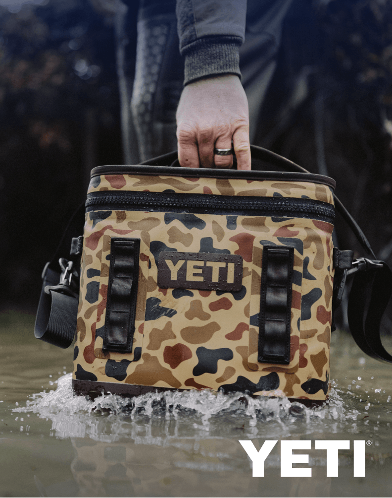 Camouflage-pattern YETI cooler bag being carried through shallow water.