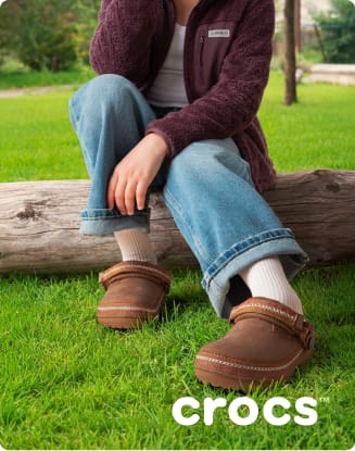 Person seated on a log outdoors wearing brown Crocs clogs, cuffed jeans, and a fleece jacket on a grassy lawn.