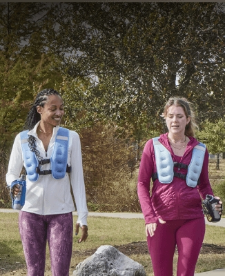 Two people walking outdoors wearing weighted vests and holding water bottles.