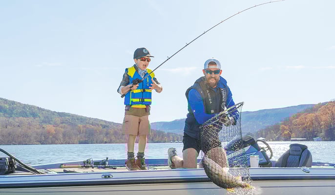 Father and son fishing fishing on a lake in a boat, wearing life vests and netting a big fish.