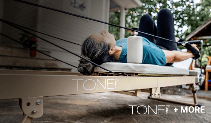 Beige pilates reformer machine outdoors with resistance bands attached, labeled TONE! With a woman shown workout using the machine.