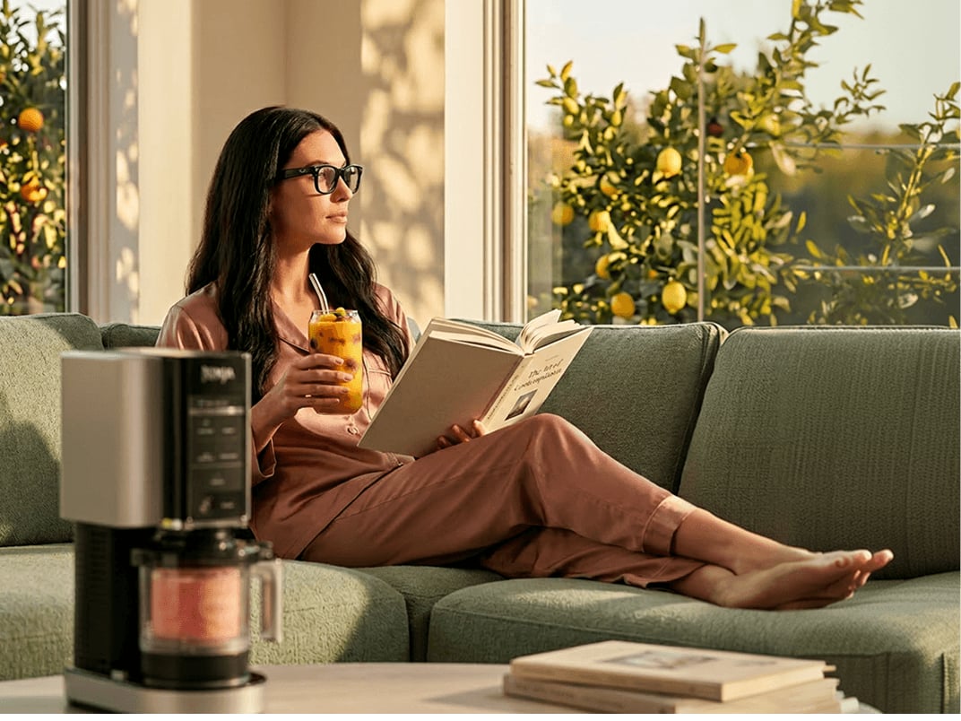Woman relaxing on a sofa indoors, enjoying a smoothie made with a Ninja Creami in a sunlit living room while wearing Ray-Ban Meta glasses.