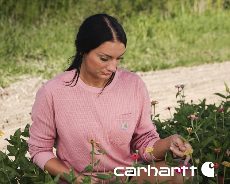 Farmer Grayce Emmick wearing a pink Carhartt sweatshirt tending plants in a garden with overlaid Carhartt logo.