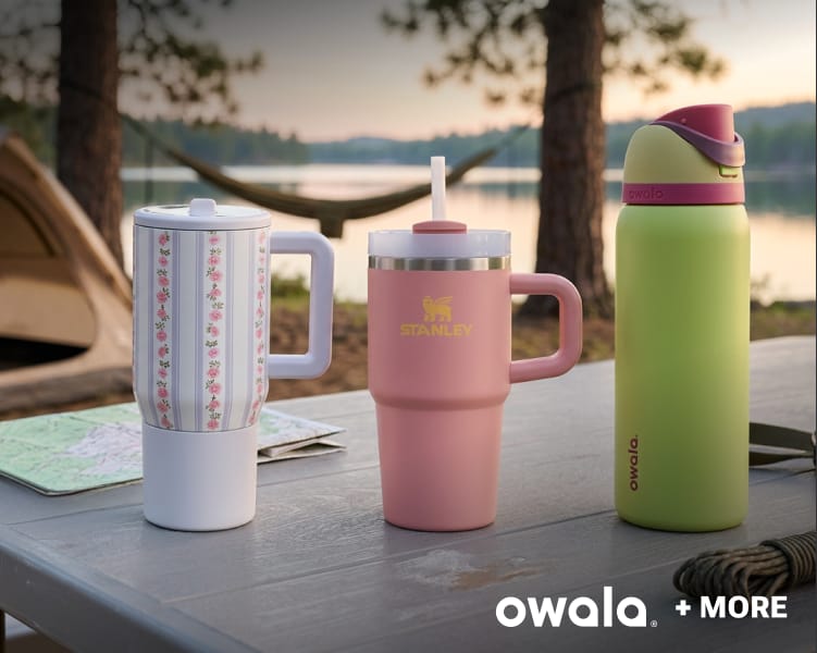 Insulated drink tumblers and water bottles displayed on a picnic table at a lakeside campsite.