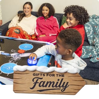Family gathered around a tabletop air hockey game in a living room. A wooden holiday sign with snow reads “Gifts for the Family.”
