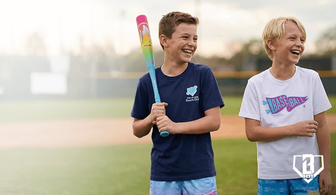 Two kids standing on a baseball field wearing colorful athletic shorts from the Swing into Spring collection from Baseball Lifestyle 101, one holding a brightly colored baseball bat.