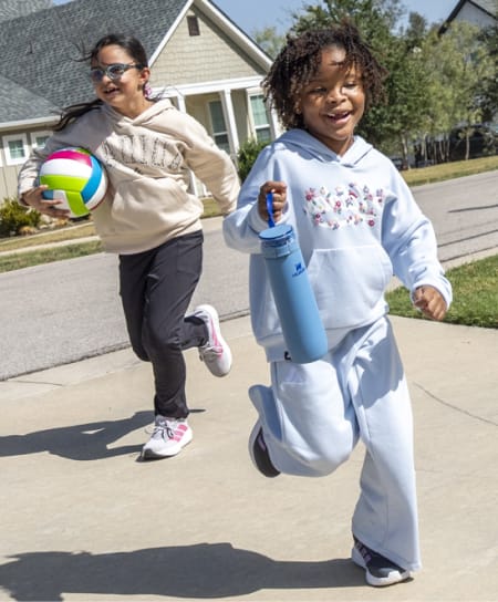 Two children running on a neighborhood sidewalk, carrying a volleyball and a water bottle while wearing casual athletic apparel.