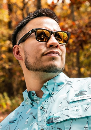 Close-up of a person wearing sunglasses and a light blue patterned button-down shirt outdoors. Trees with autumn-colored leaves fill the background.