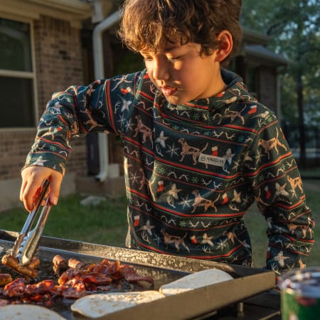 Child wearing a Magellan Outdoors holiday sweater uses grilling tongs at an outdoor griddle.
