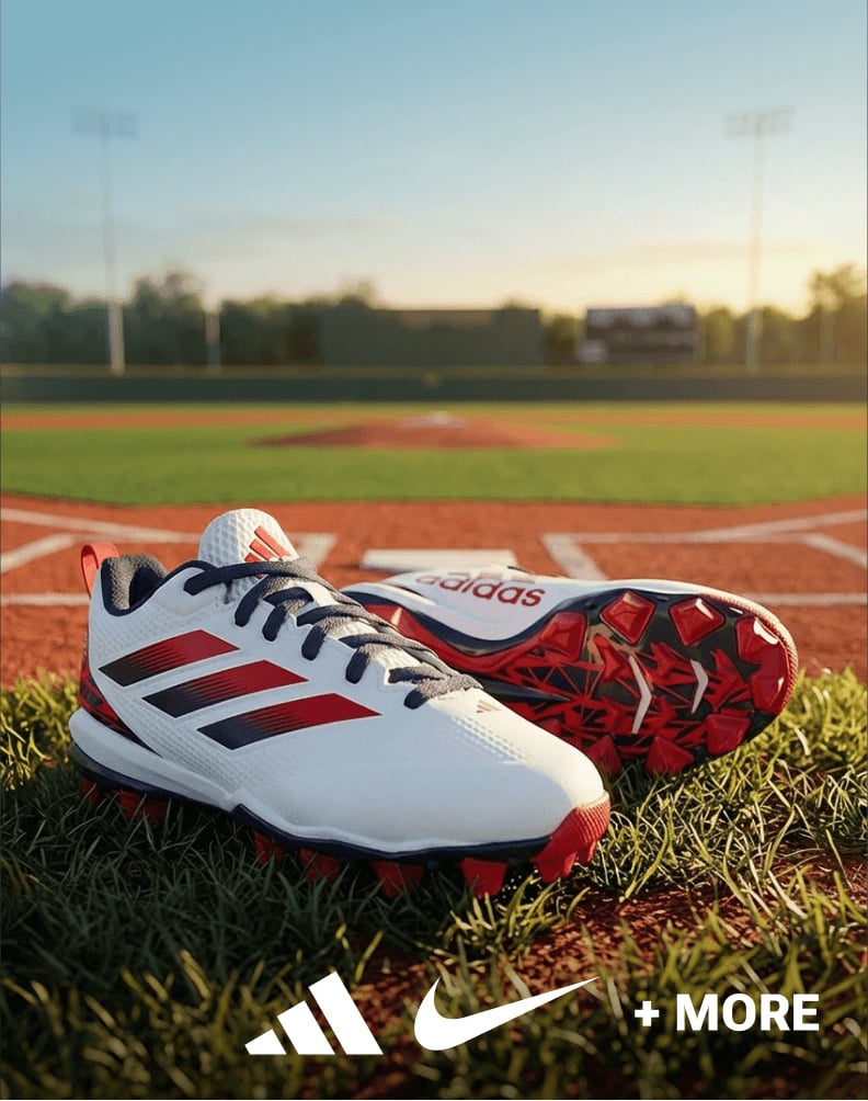 A pair of white adidas baseball cleats with red and navy accents placed on grass near a baseball field.