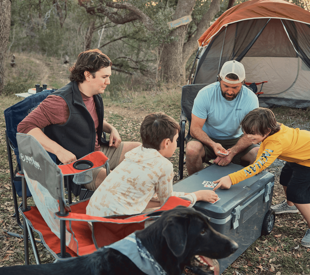 Group of people gathered at a campsite near a tent, seated around a portable table with outdoor gear.